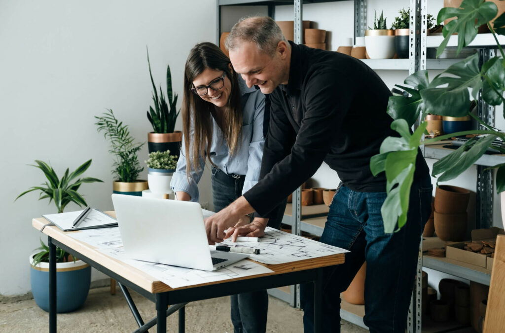 A man and woman looking over laptop and building plans on desk